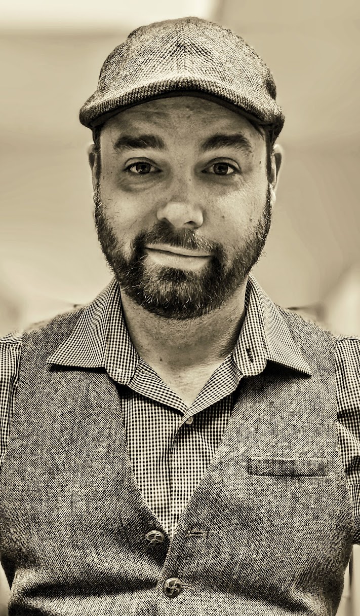 Man with a beard wearing a flat cap and a vest, smiling, in an indoor setting.