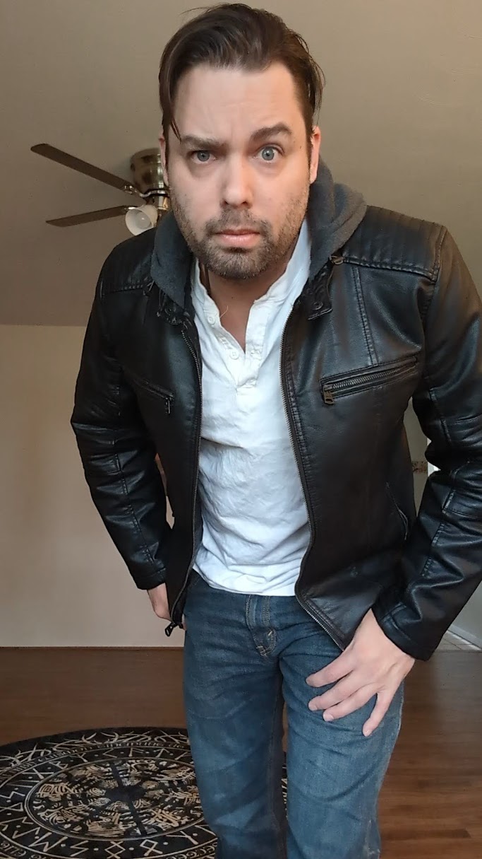 Man with dark hair and beard wearing a black leather jacket and white shirt, standing indoors with a ceiling fan in background.
