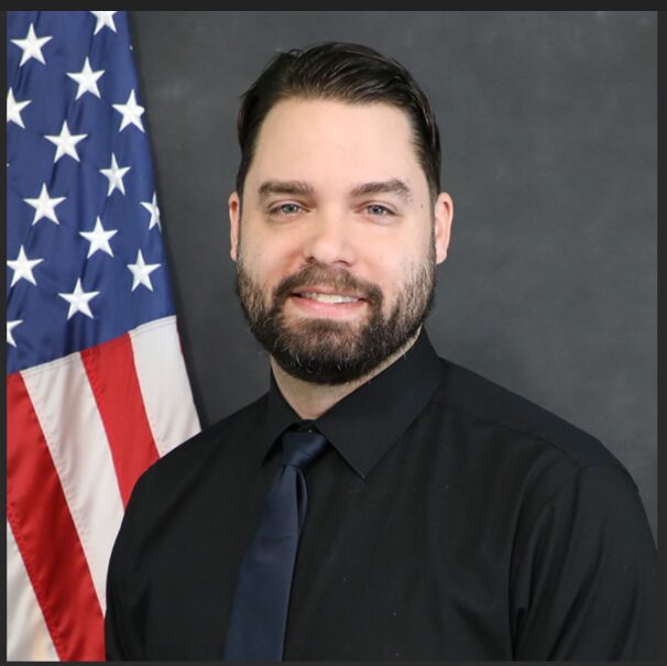 Man with dark hair and beard wearing a black shirt and tie, standing in front of an American flag, smiling.