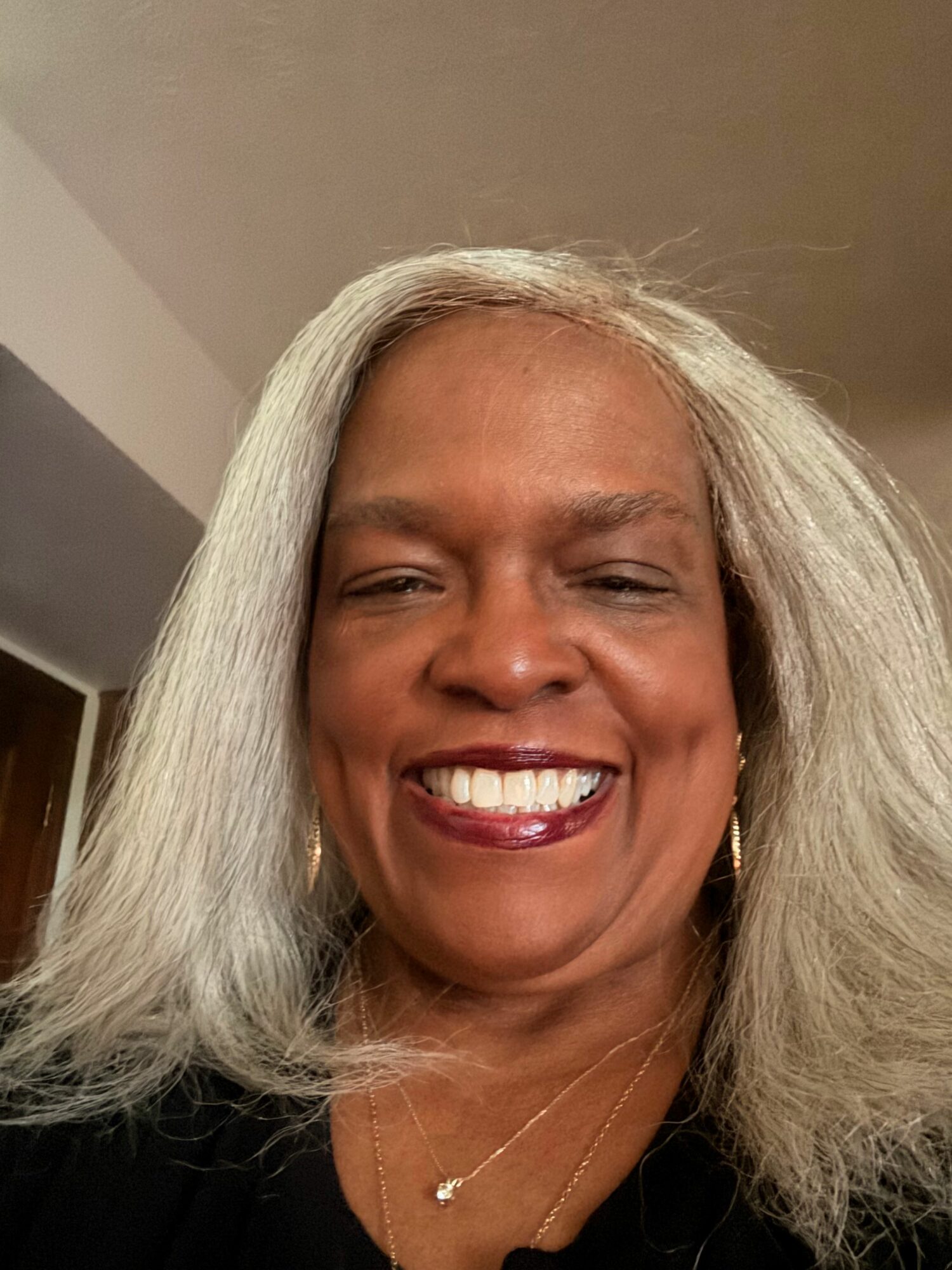 Smiling woman with long gray hair, wearing a black top and a necklace, facing the camera.