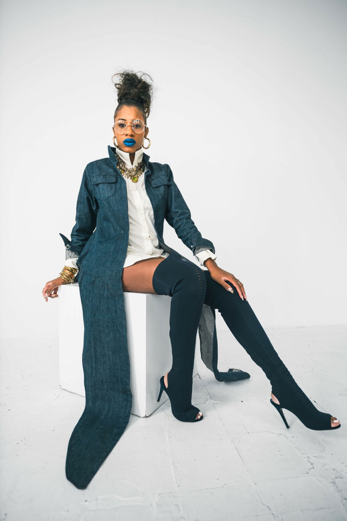 Woman sitting on a white box, wearing a denim jacket, white shirt, black thigh-high boots, and jewelry, against a plain white background.