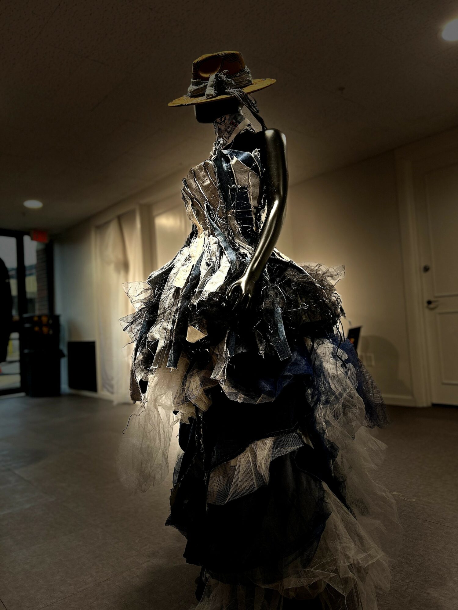 A tall, artistic mannequin dressed in layered black and white fabric, wearing a wide-brimmed hat, in an indoor setting.