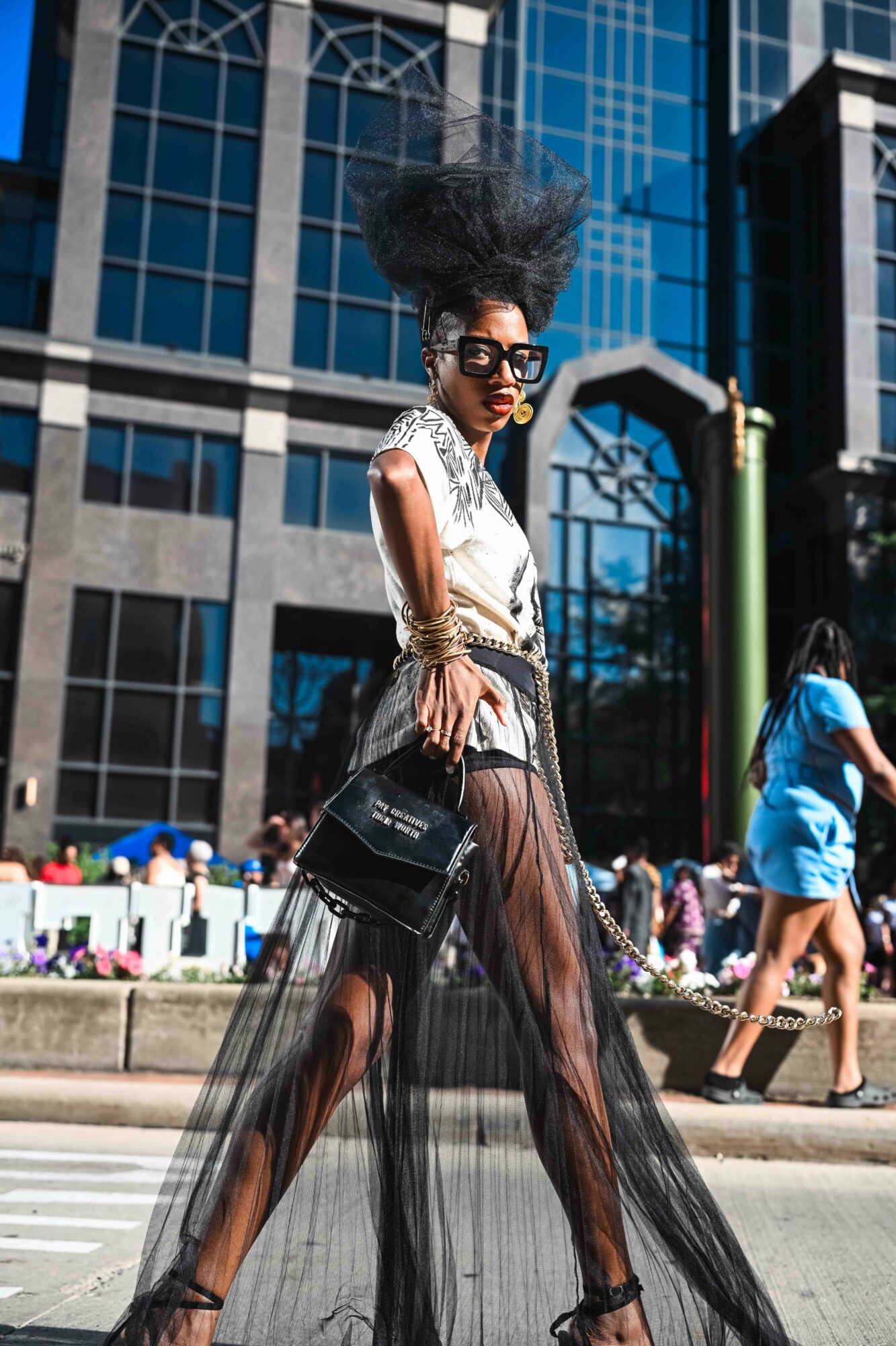 Fashionably dressed woman with large hair and sunglasses walking on city street, tall buildings in background.