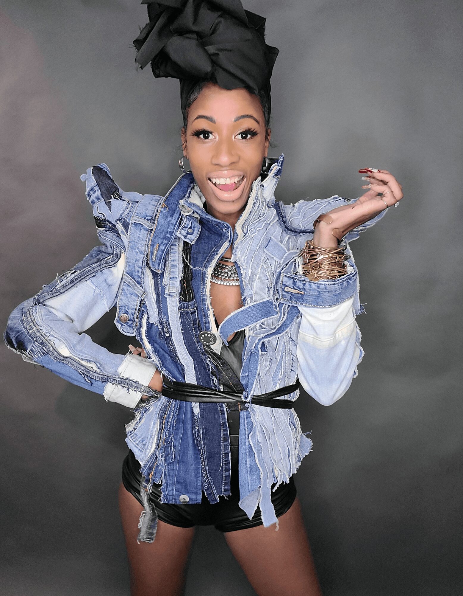 Woman with styled hair, wearing a denim jacket with ruffled shoulders, smiling, against a gray background.