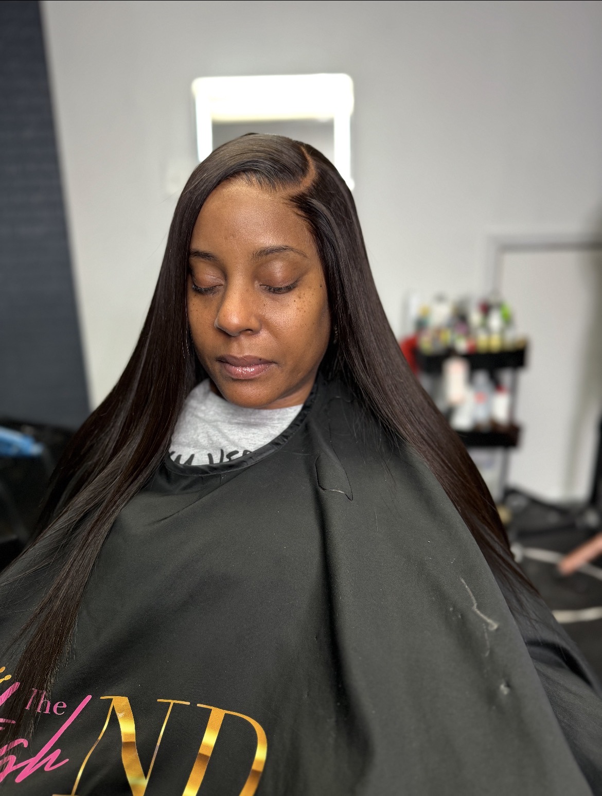 Woman with long straight hair sitting in a salon chair, wearing a black cape, with a mirror and salon supplies in background.