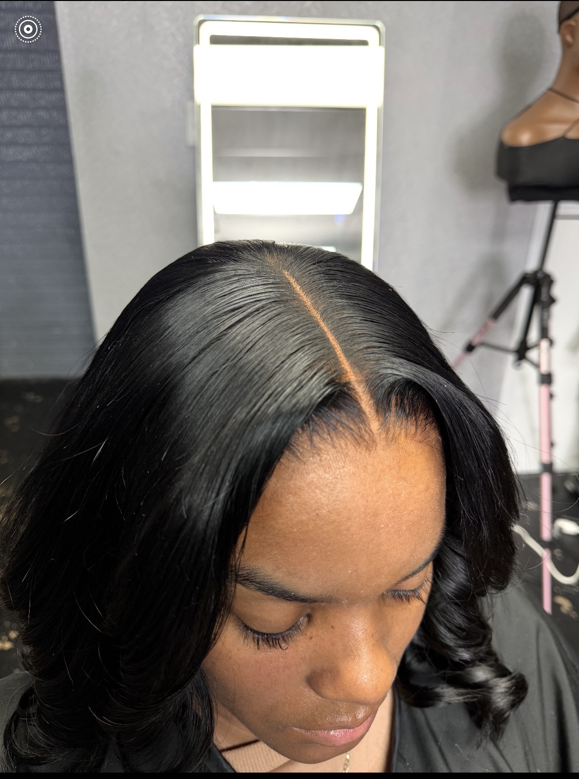 Top view of a woman with long, shiny black hair styled in loose waves, looking downward, with a white chair in the background.