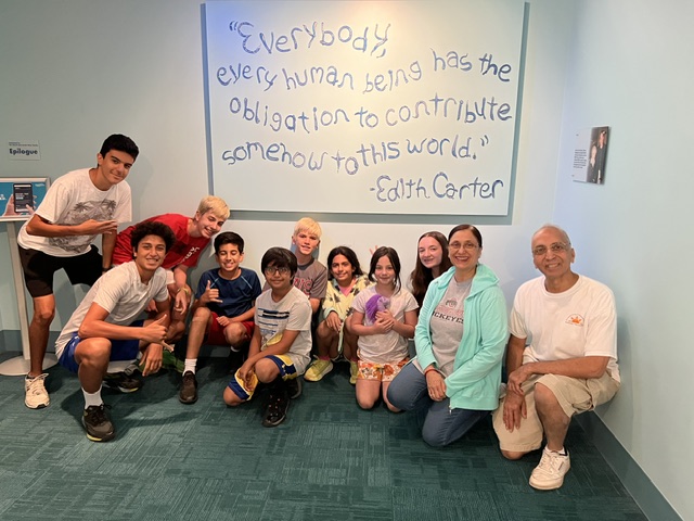 Group of nine people, including children and adults, posing in front of a whiteboard with a quote, in a room with light blue walls.