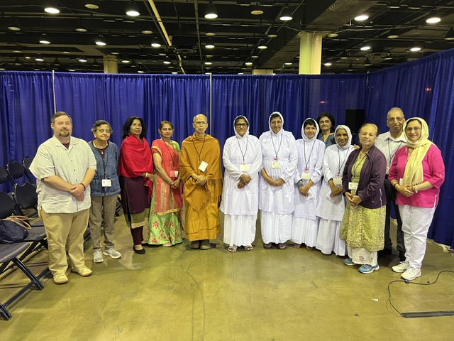 Group of people standing in front of blue curtain, some in traditional and others in casual clothing, indoors.