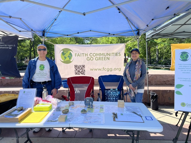 Two people stand behind a table at an outdoor event under a blue canopy, with a banner reading 'Faith Communities Go Green' behind them.
