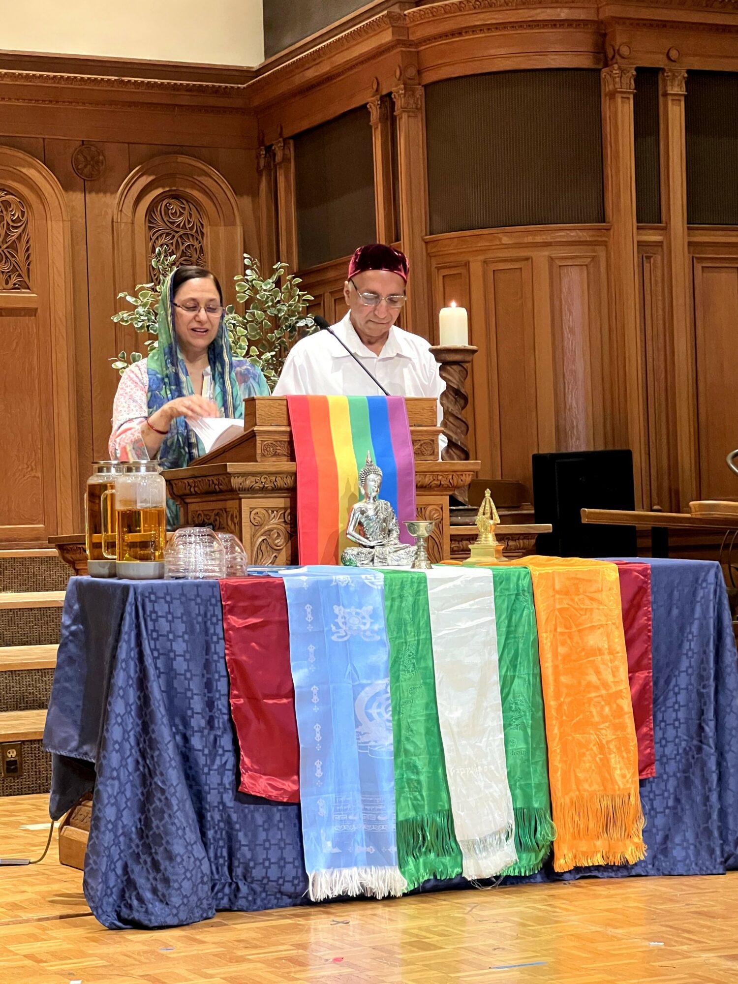 Two people stand behind a table with rainbow flags, candles, and a glass of beer in a wood-paneled room.