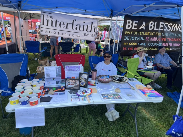 Table with religious items and photos under a blue canopy at an outdoor event, with people seated behind.