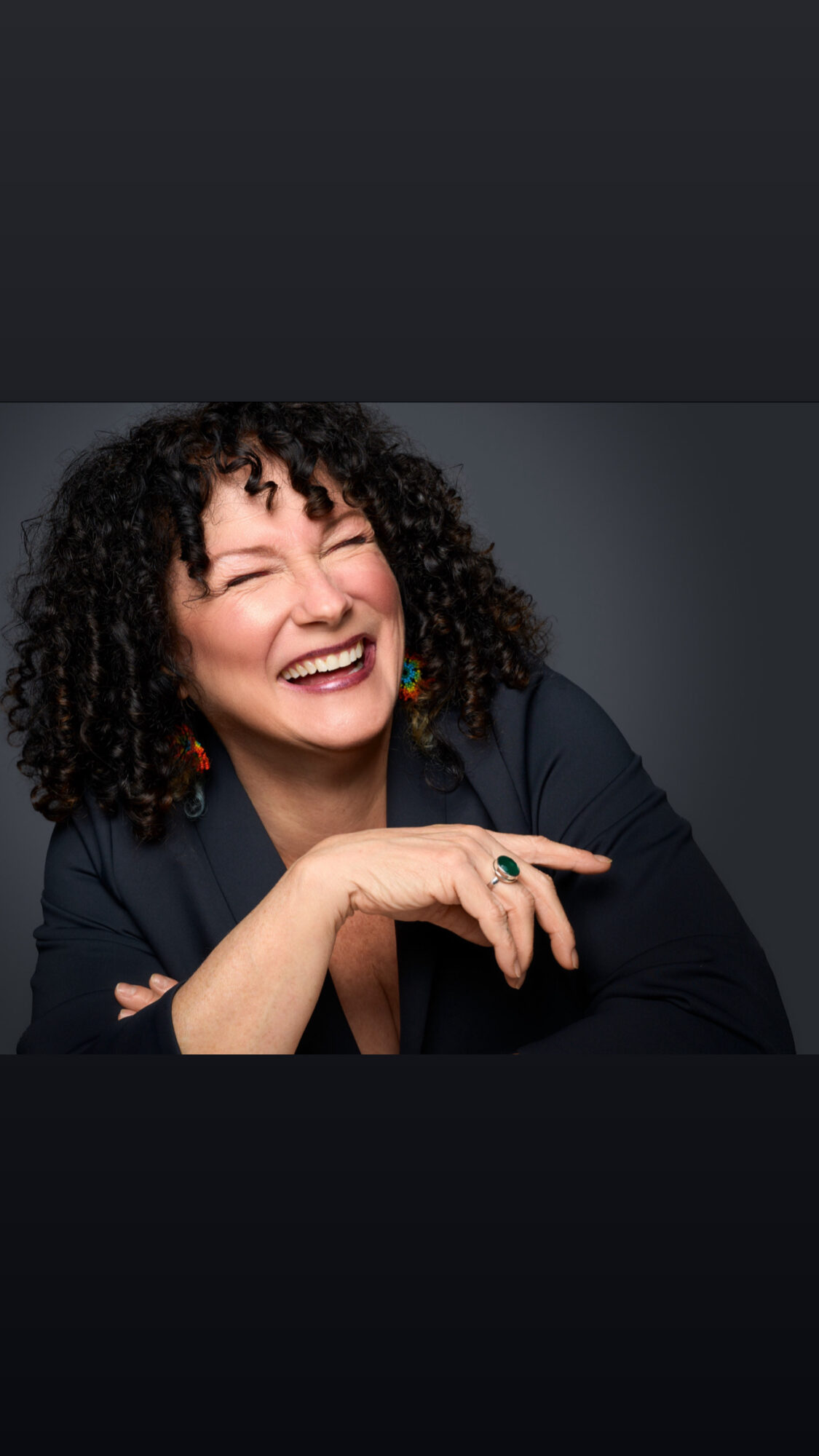 Woman with curly hair smiling, wearing a dark blazer, resting her chin on her hand, against a dark background.
