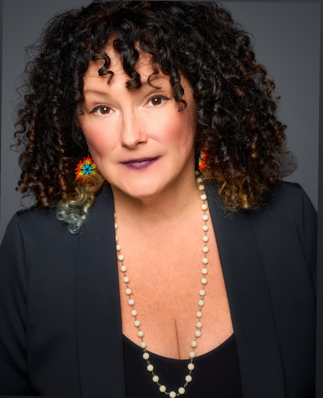 Woman with curly hair wearing earrings, a pearl necklace, and a dark blazer, looking at the camera.