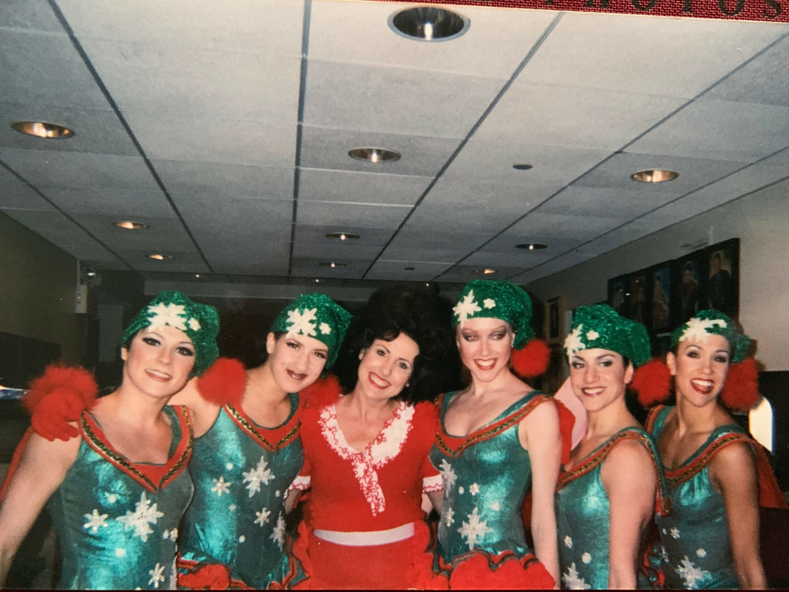 Six women in festive costumes and a woman in a red dress pose together indoors, smiling at the camera.