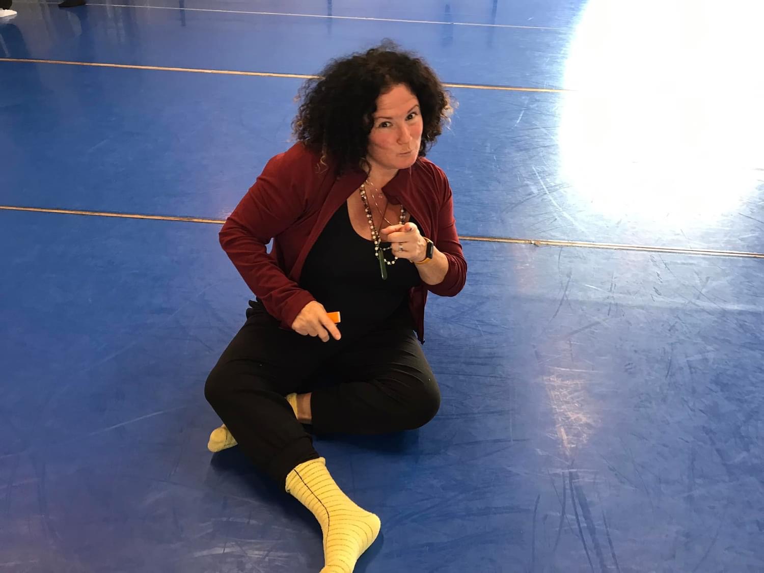 Woman with curly hair kneeling on a blue floor, wearing a red jacket, black pants, and yellow socks, holding a phone.