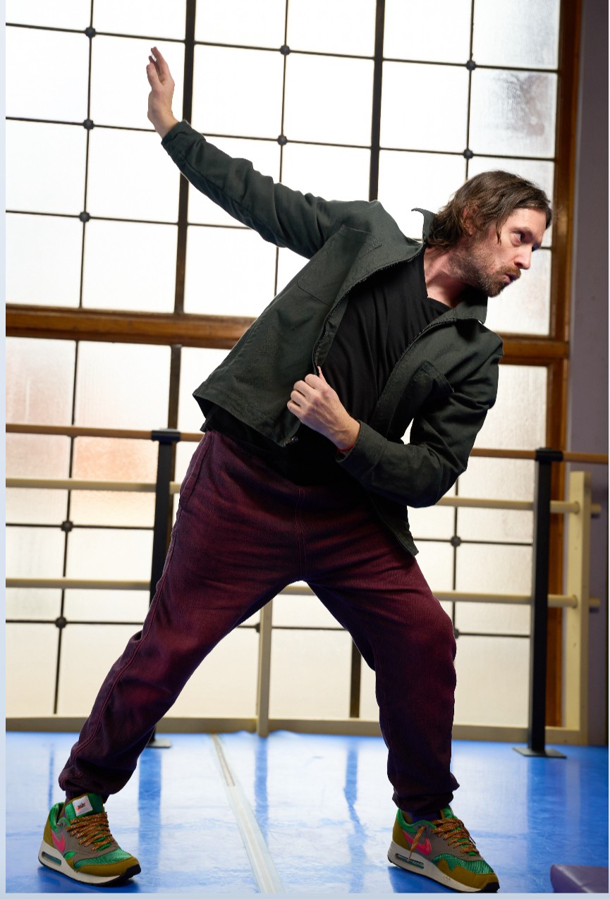 Man in black jacket and maroon pants strikes a dance pose indoors with a grid window background.