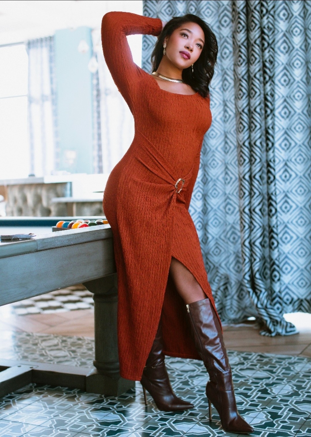 Woman in a long red dress and knee-high boots poses indoors near a window and blue patterned curtains.