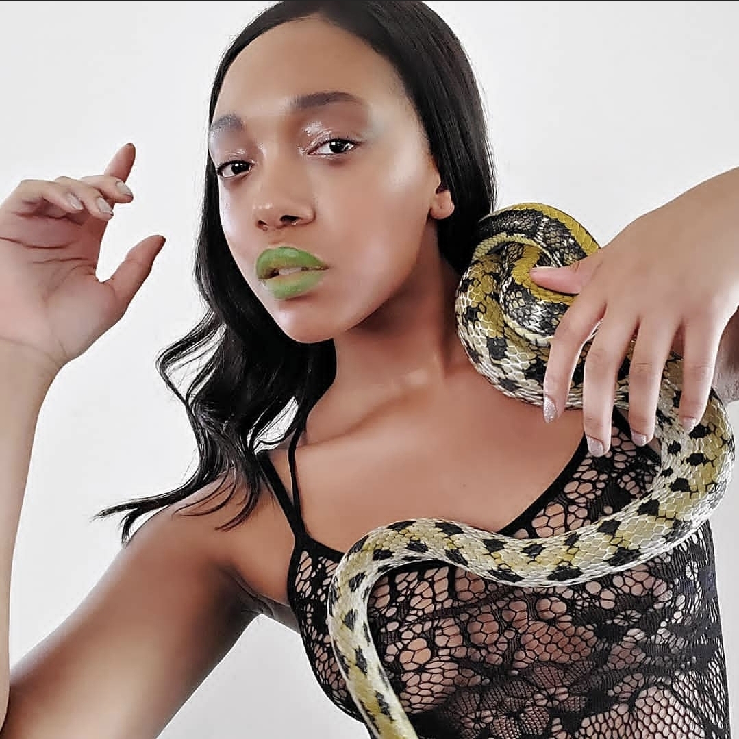 Woman with black hair and green lipstick holding a snake on her shoulder, wearing a black lace top, against a white background.