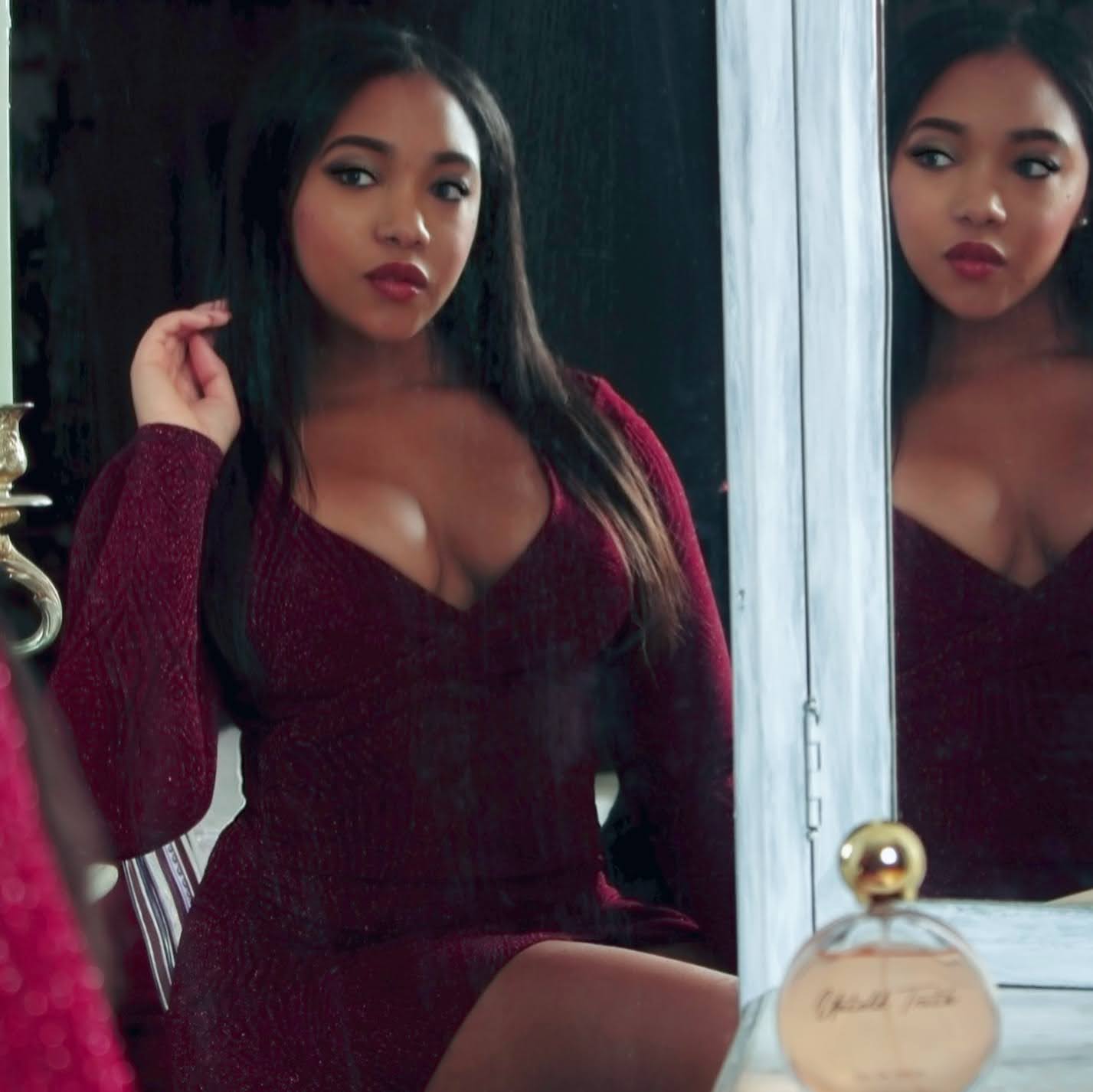 Woman with long dark hair in a deep red dress looking at mirror, hand near face, reflection visible, indoor setting