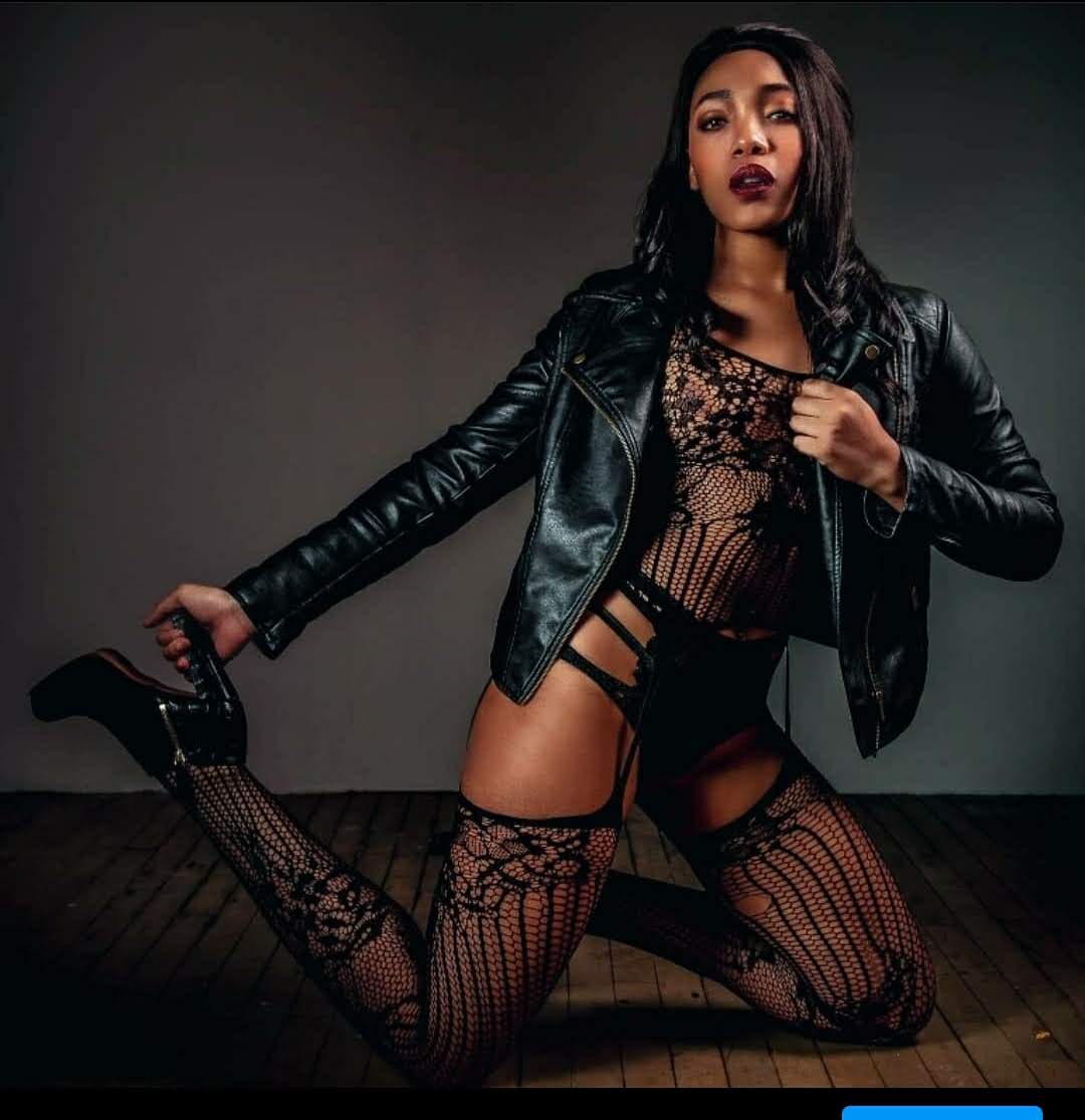 Woman in black leather jacket and fishnet stockings kneels on floor, holding a high-heeled shoe, against dark background.