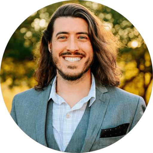 Man with long hair and beard smiling outdoors, wearing a suit and light shirt, with trees in background.