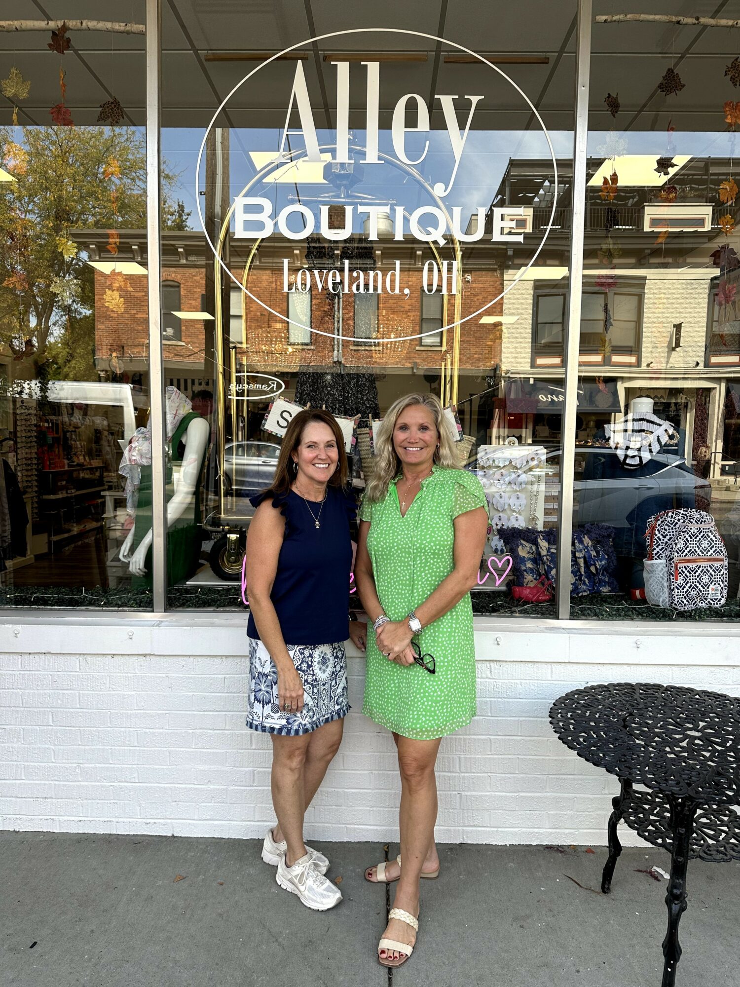 Two women stand outside a boutique shop window, smiling, with buildings and cars reflected in the glass.