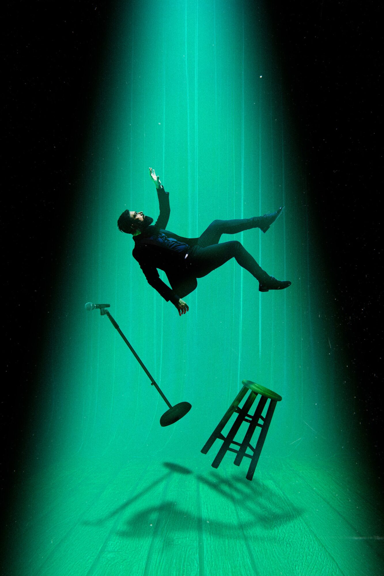 Person floating in a spotlight with a stool and microphone below, dark background, illuminated by teal light.