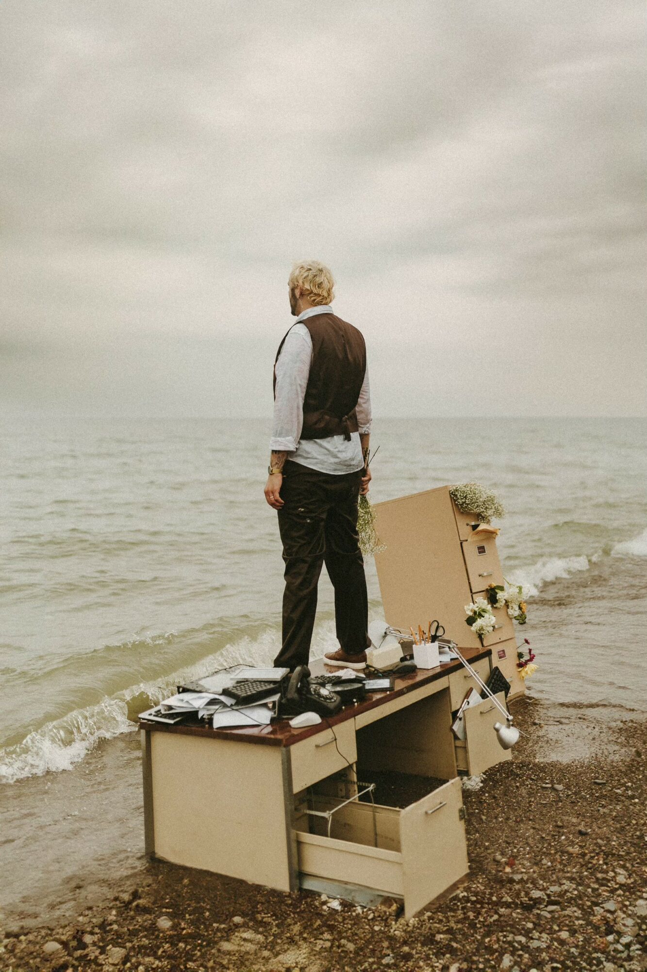 Person standing on a desk at the beach, facing the ocean, with cardboard boxes and flowers nearby.