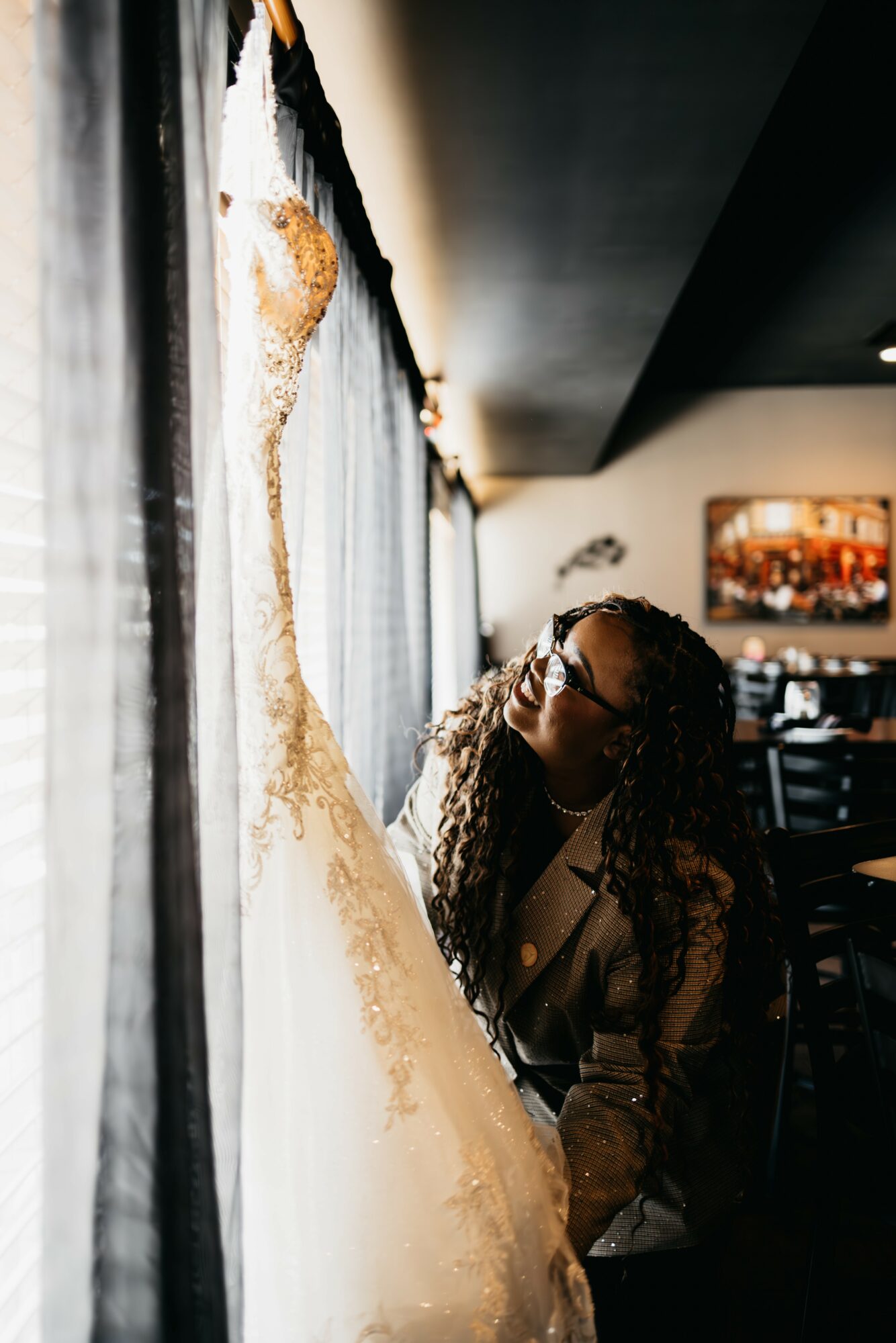 Woman with curly hair and glasses looks at a wedding dress hanging on a wall, in a dimly lit room.