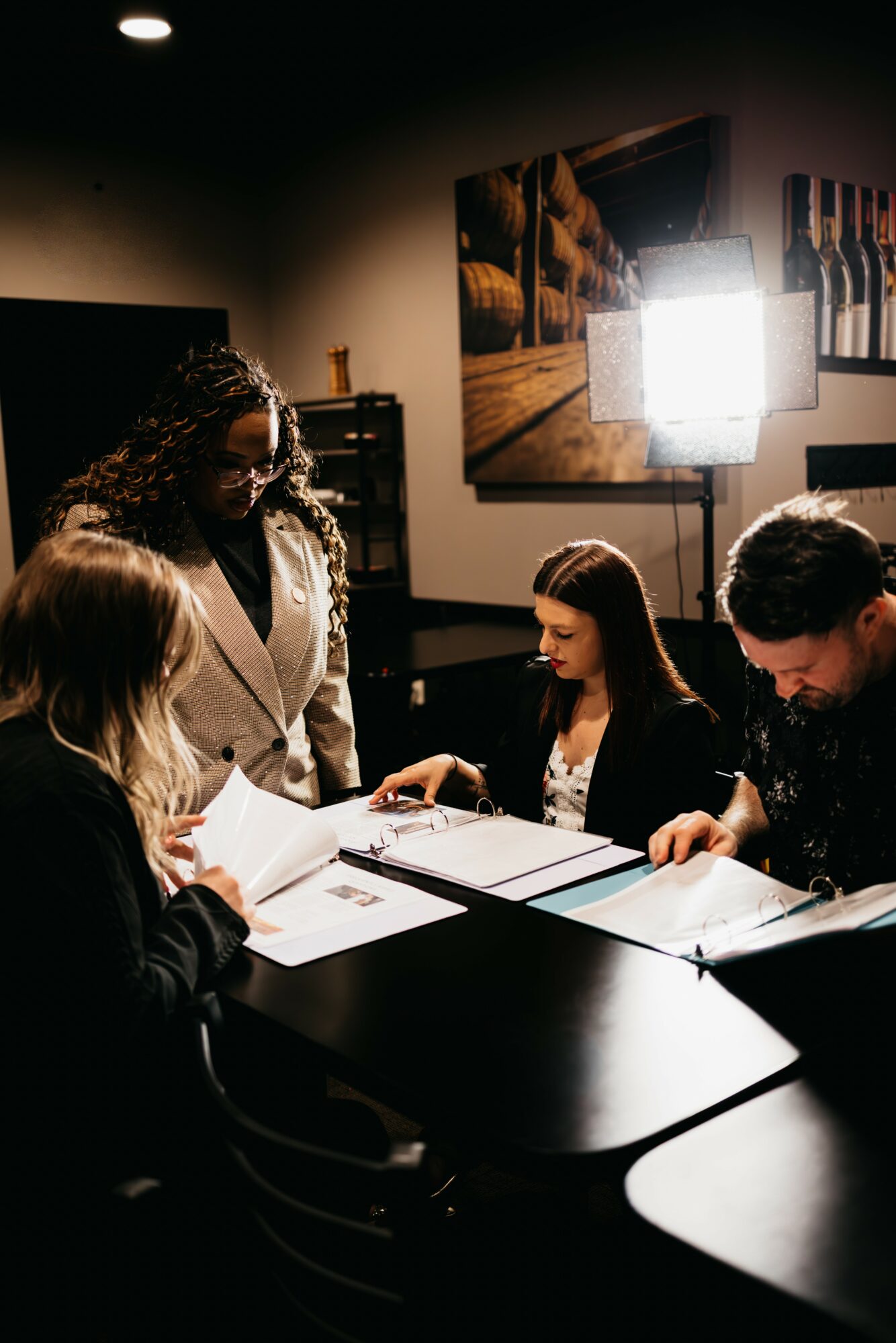 Four people sit around a table in a dimly lit room, engaged in discussion with documents and notebooks, with a bright light and artwork on the wall.