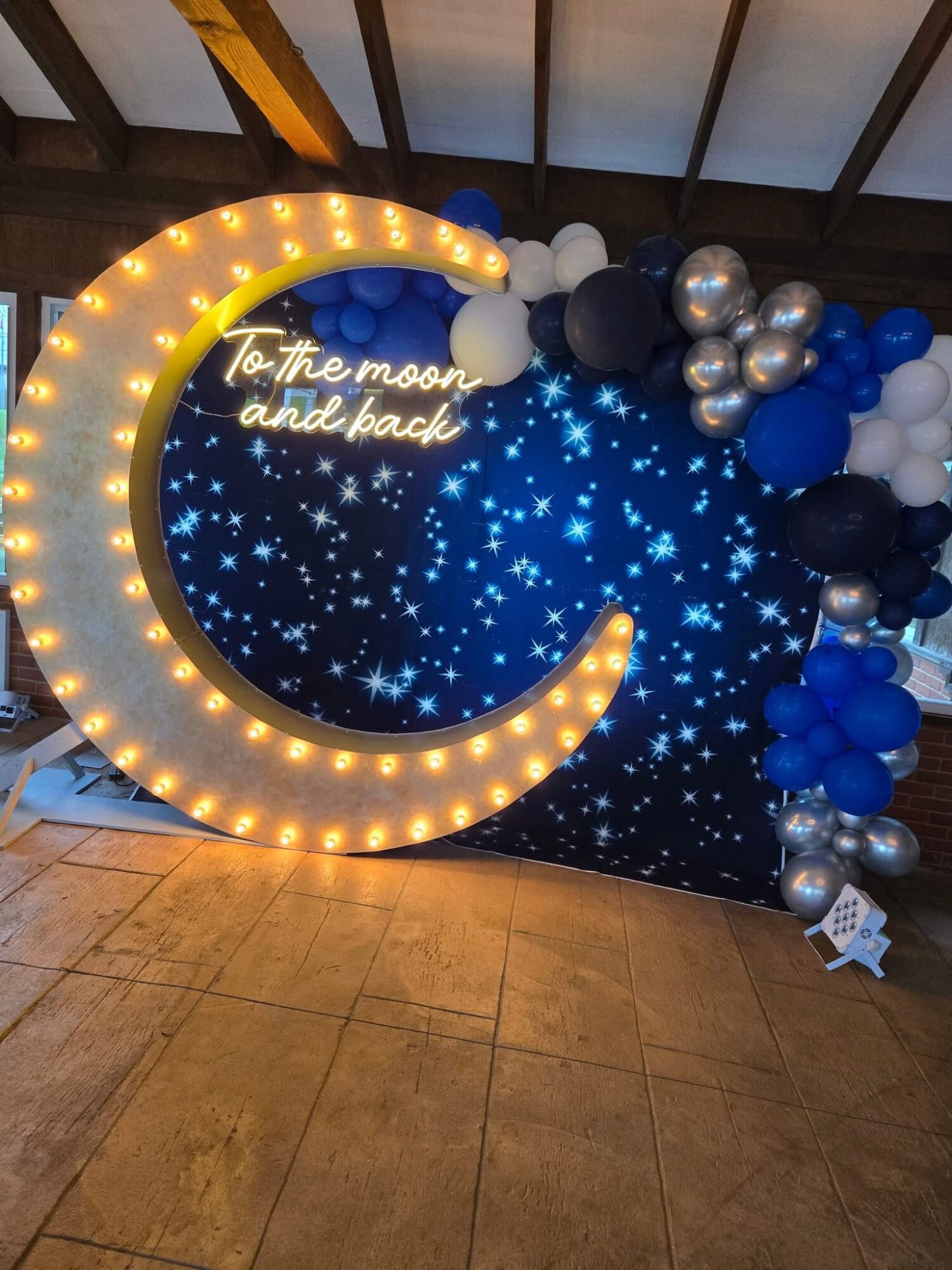 Decorative moon with lights, starry backdrop, and balloons, with a neon sign reading 'To the moon and back'.