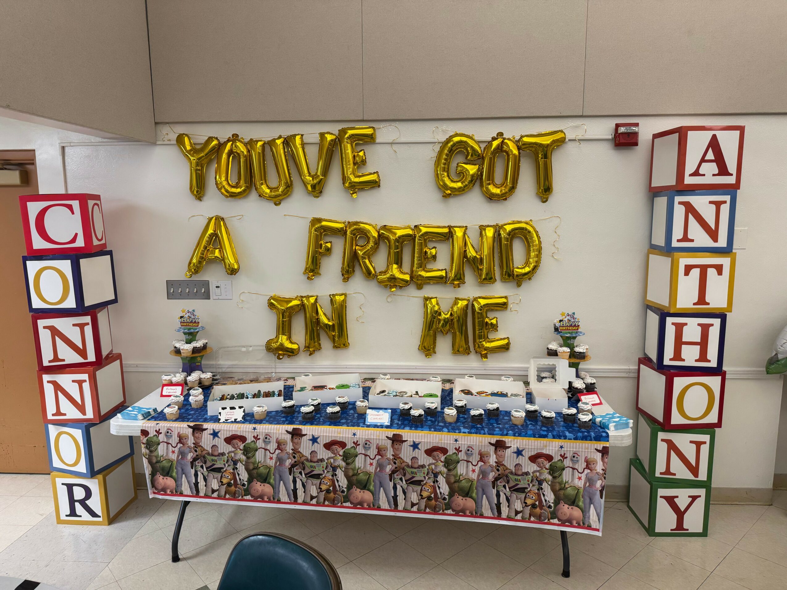 Decorative wall with gold balloons spelling 'YOU'VE GOT A FRIEND IN ME' and colorful boxes spelling 'CONNOR' and 'ANTHONY' on sides, with a table and toys below.