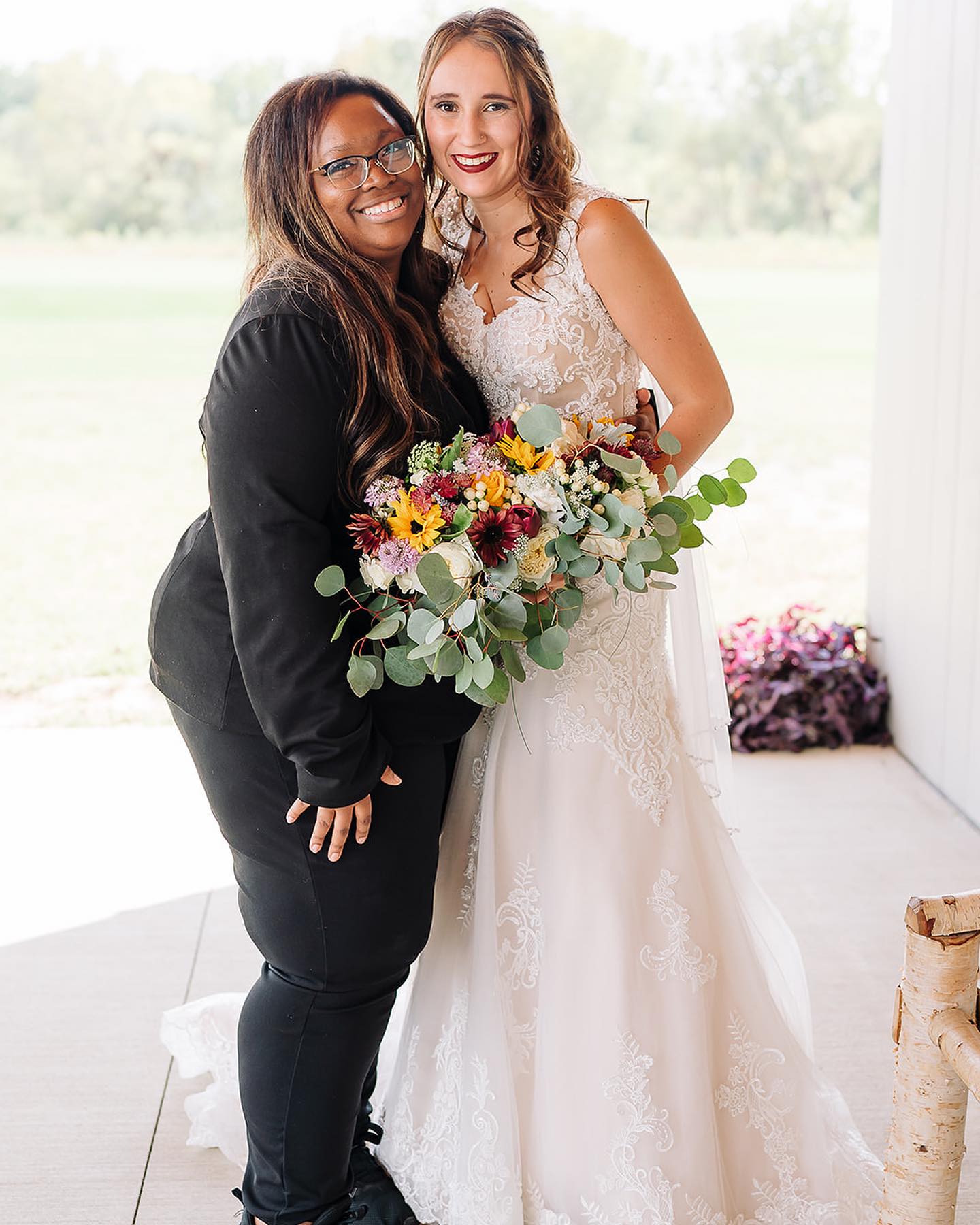 Two women, one in a wedding dress and the other in black, smiling and holding a bouquet of flowers indoors.