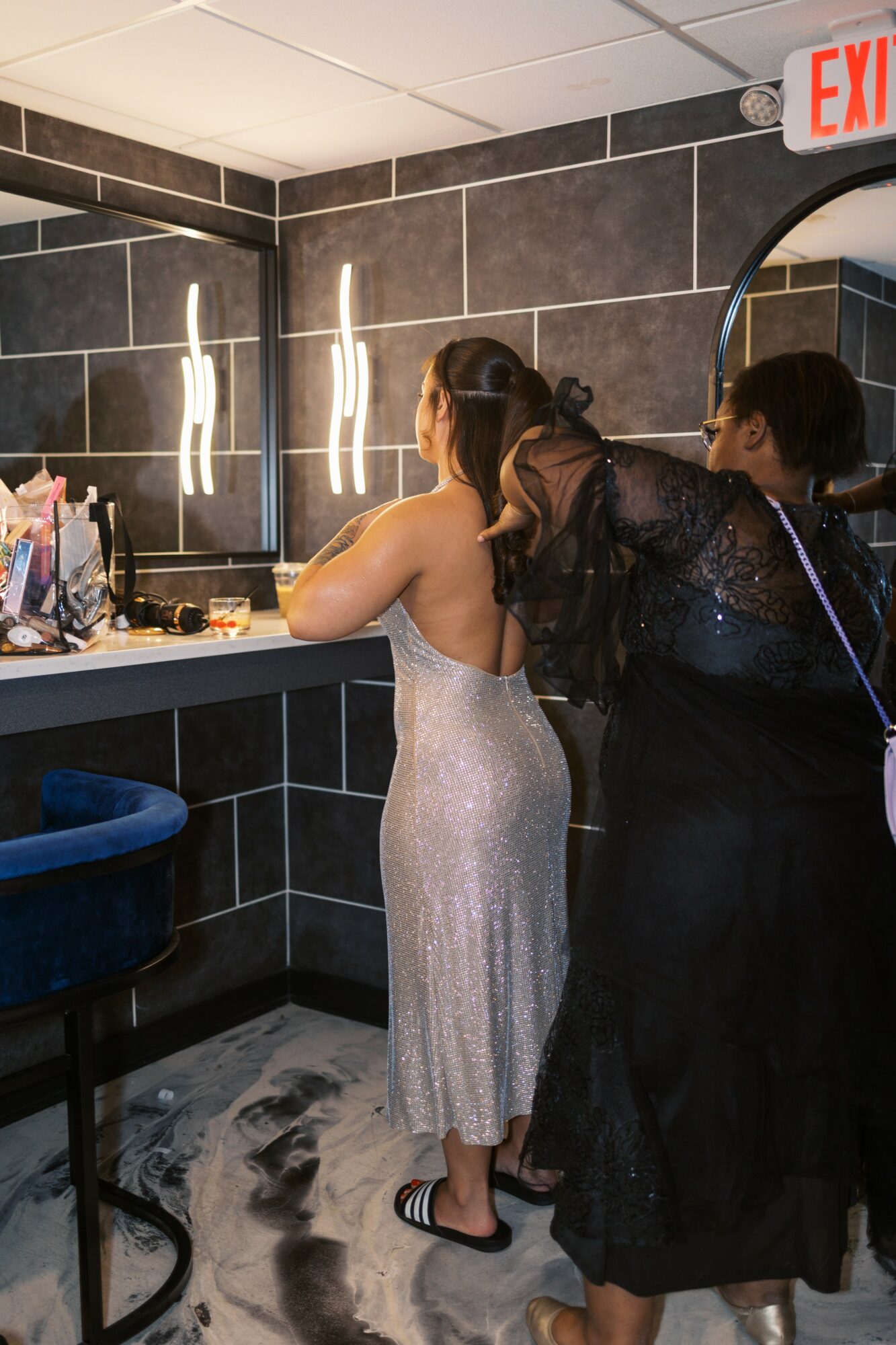Two women in elegant dresses stand at a vanity mirror, preparing for an event in a modern bathroom with black tiles.