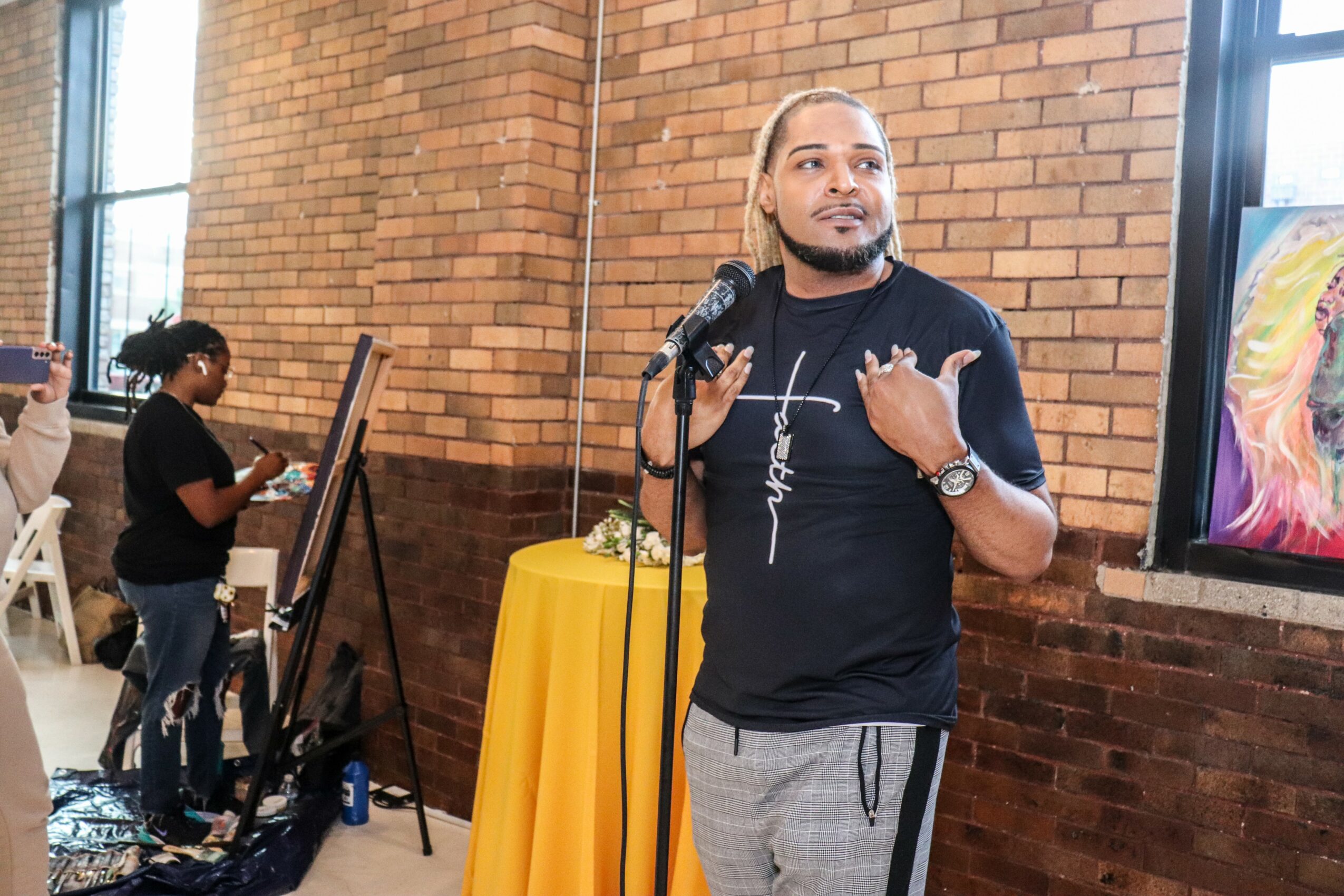 Man speaking into microphone at event, brick wall background, woman in black shirt and glasses in background, yellow table nearby.
