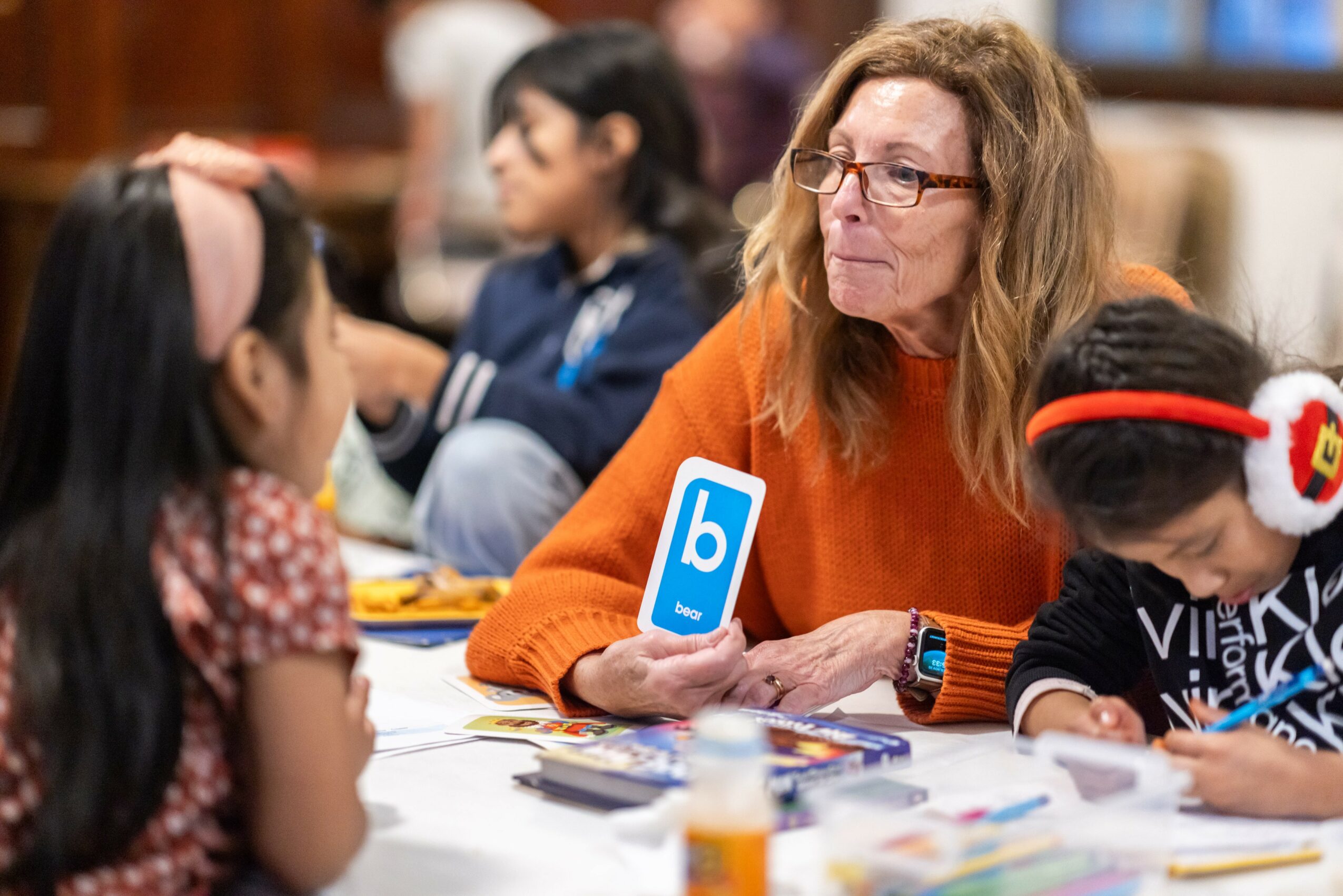 Adult woman with glasses and long hair sitting at a table with children, holding a blue card with a white letter 'b'.