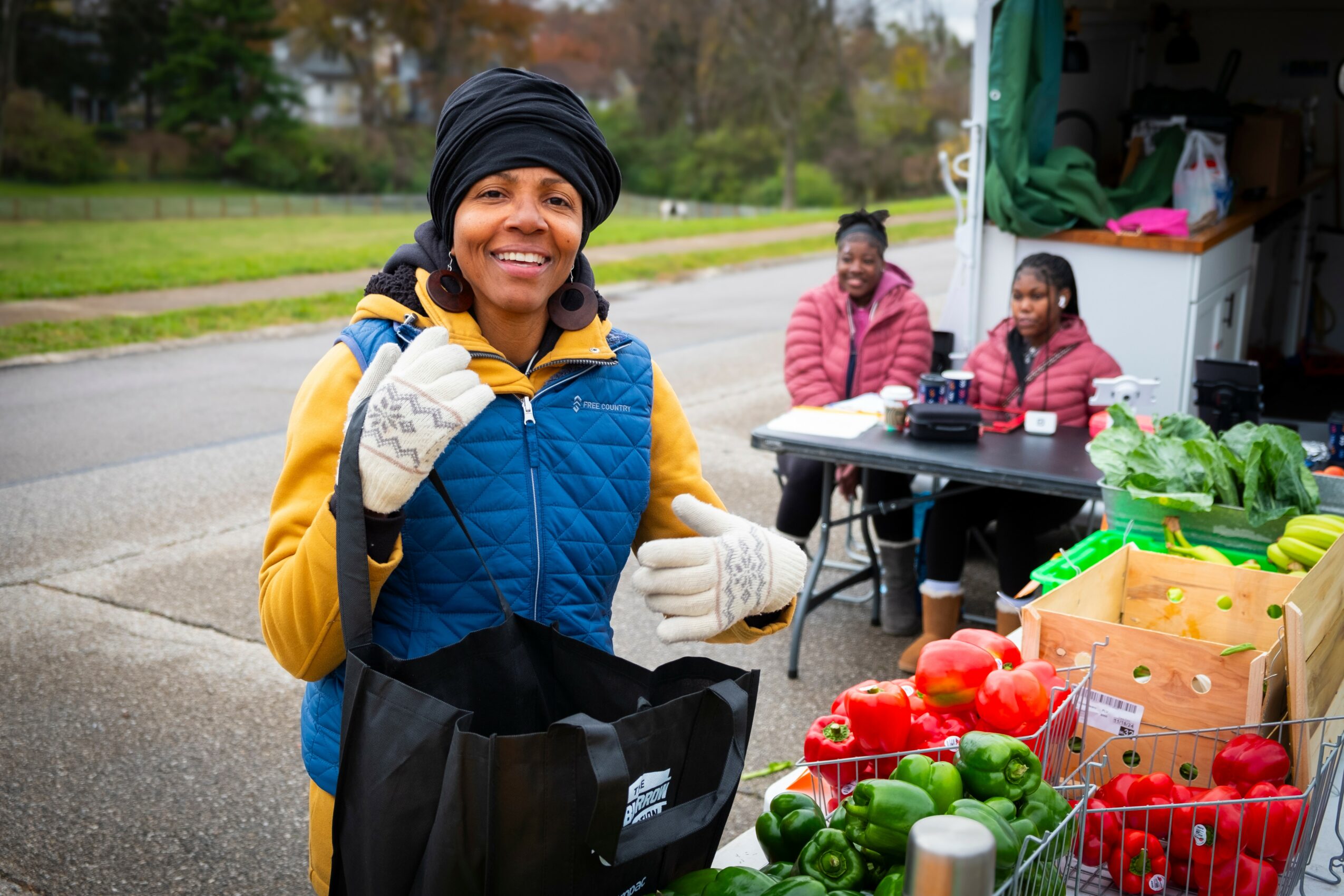Smiling woman in a blue vest and yellow hoodie at outdoor market with fresh vegetables, with two women in the background near a truck.