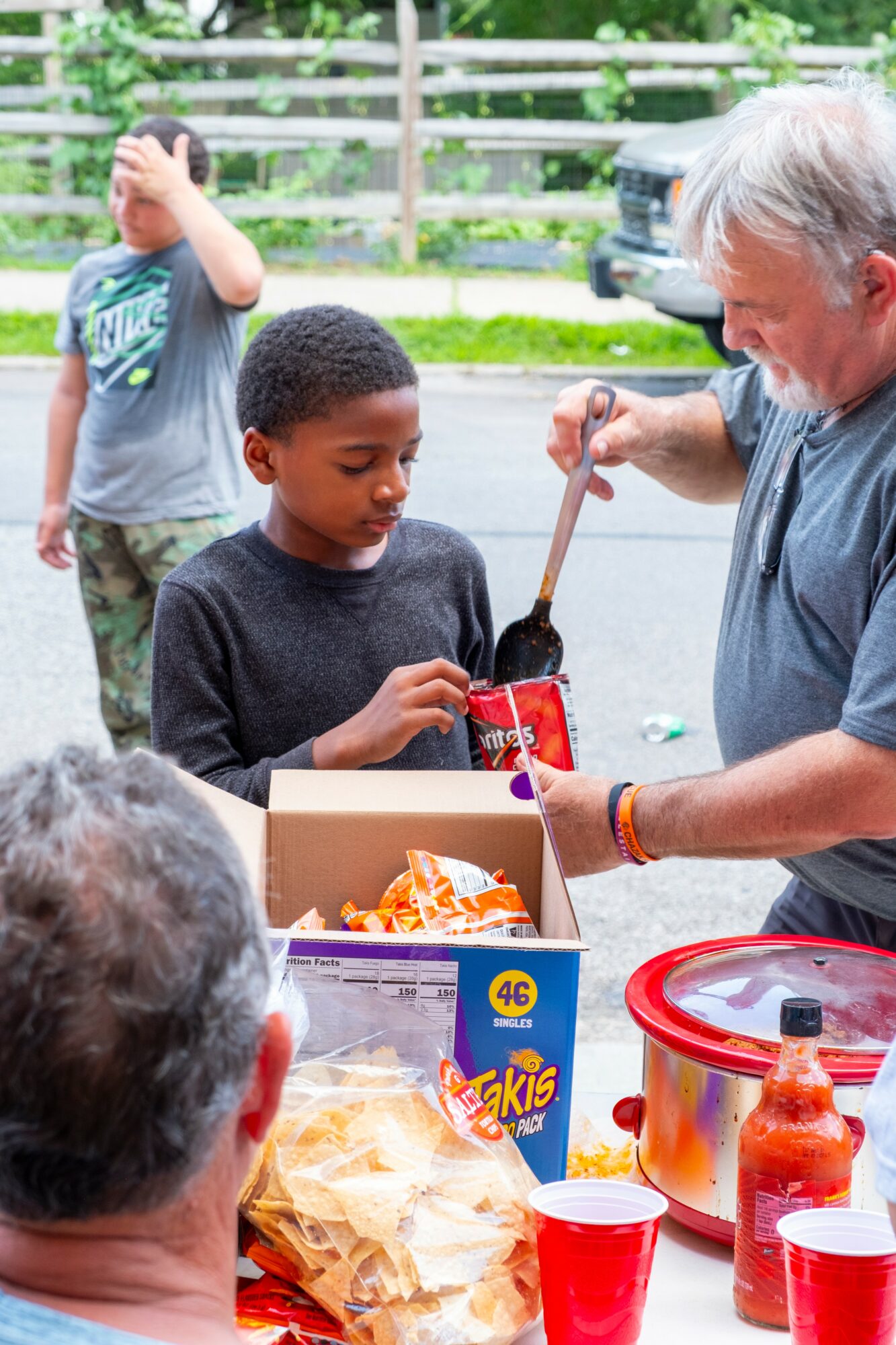 A young boy receives food from an older man at an outdoor gathering, with another child in the background and food on the table.