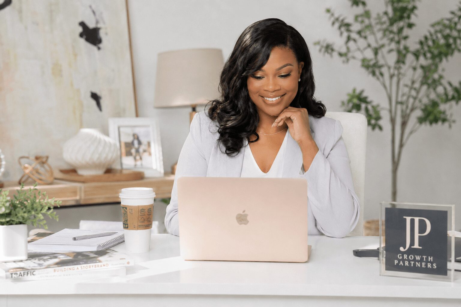 Smiling woman with dark hair working on a laptop at a desk in a bright room, with plants and decor in background.