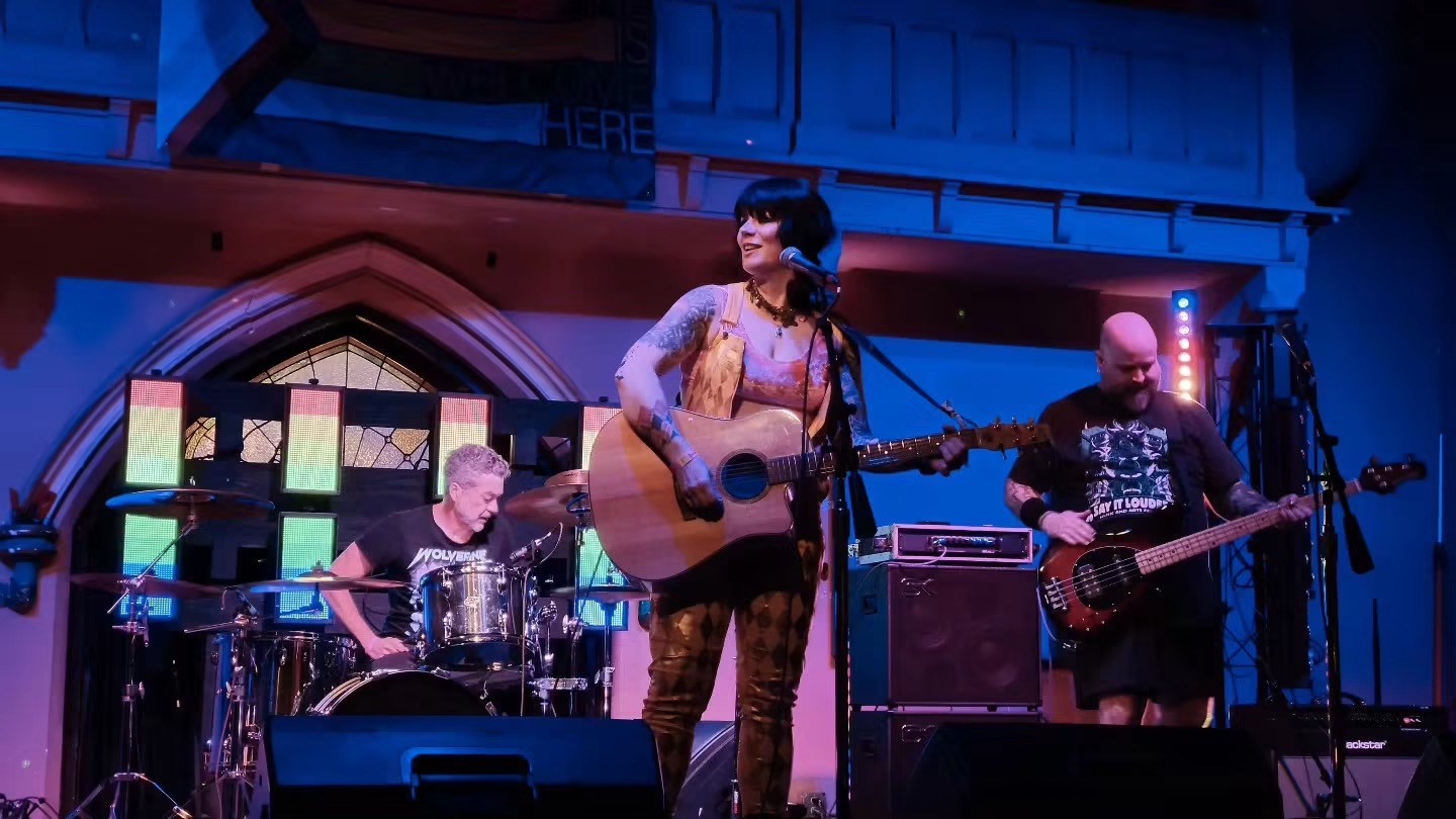 Three musicians perform on stage with colorful lighting and a decorative background, one playing guitar, others on drums and bass.