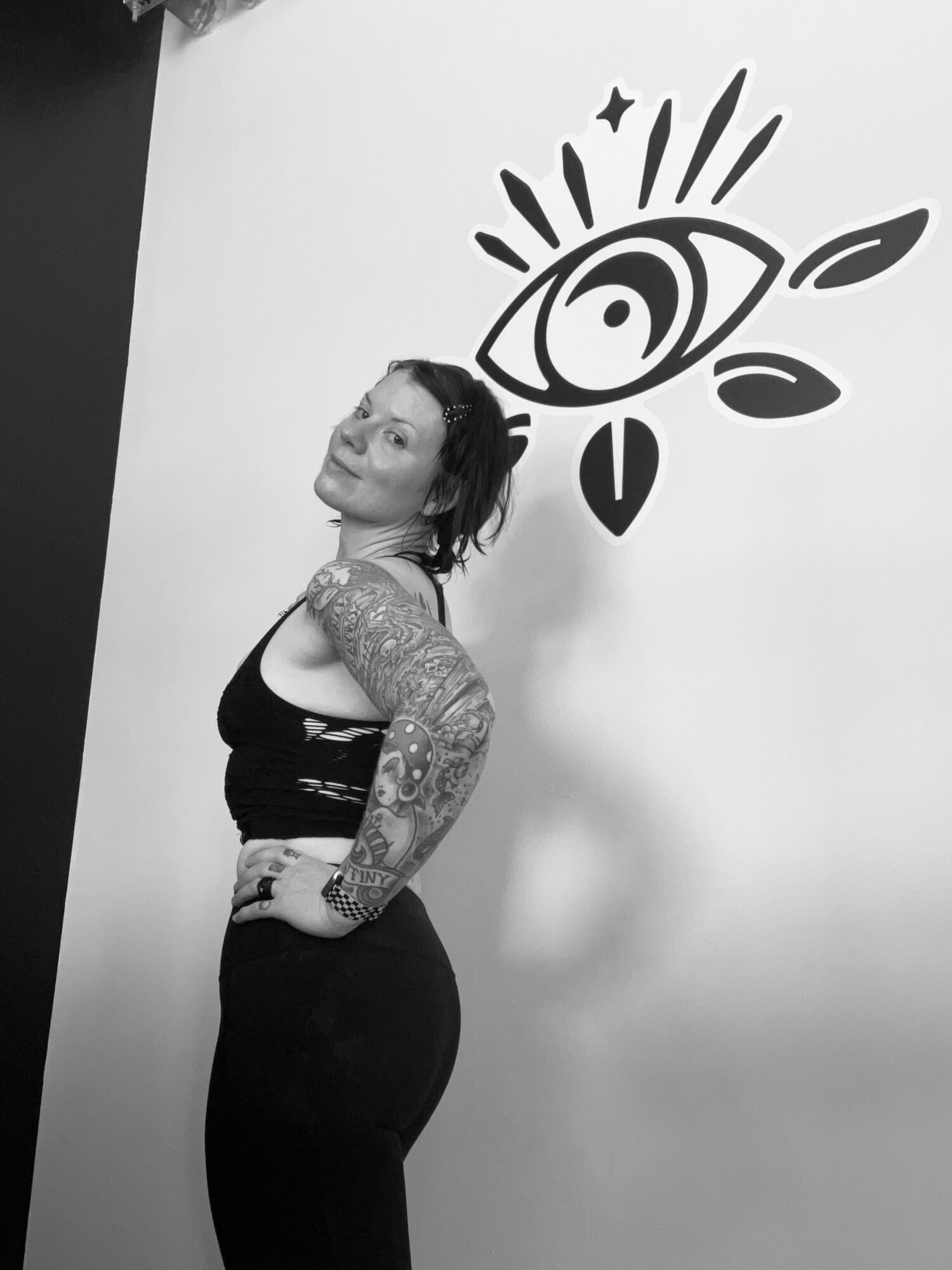 Woman with tattoos on arm, standing sideways, looking at camera, in front of a wall with eye and star design.