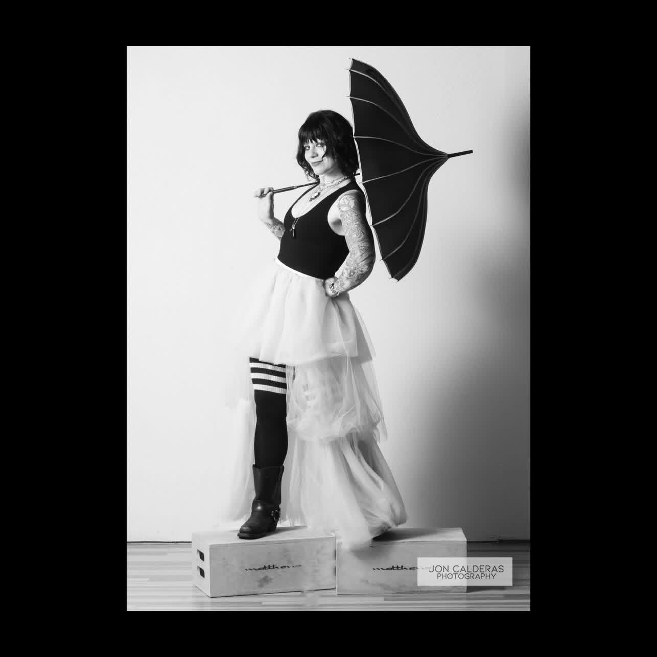 Person holding an umbrella, standing on a platform, wearing a dress and striped socks, in a studio setting.