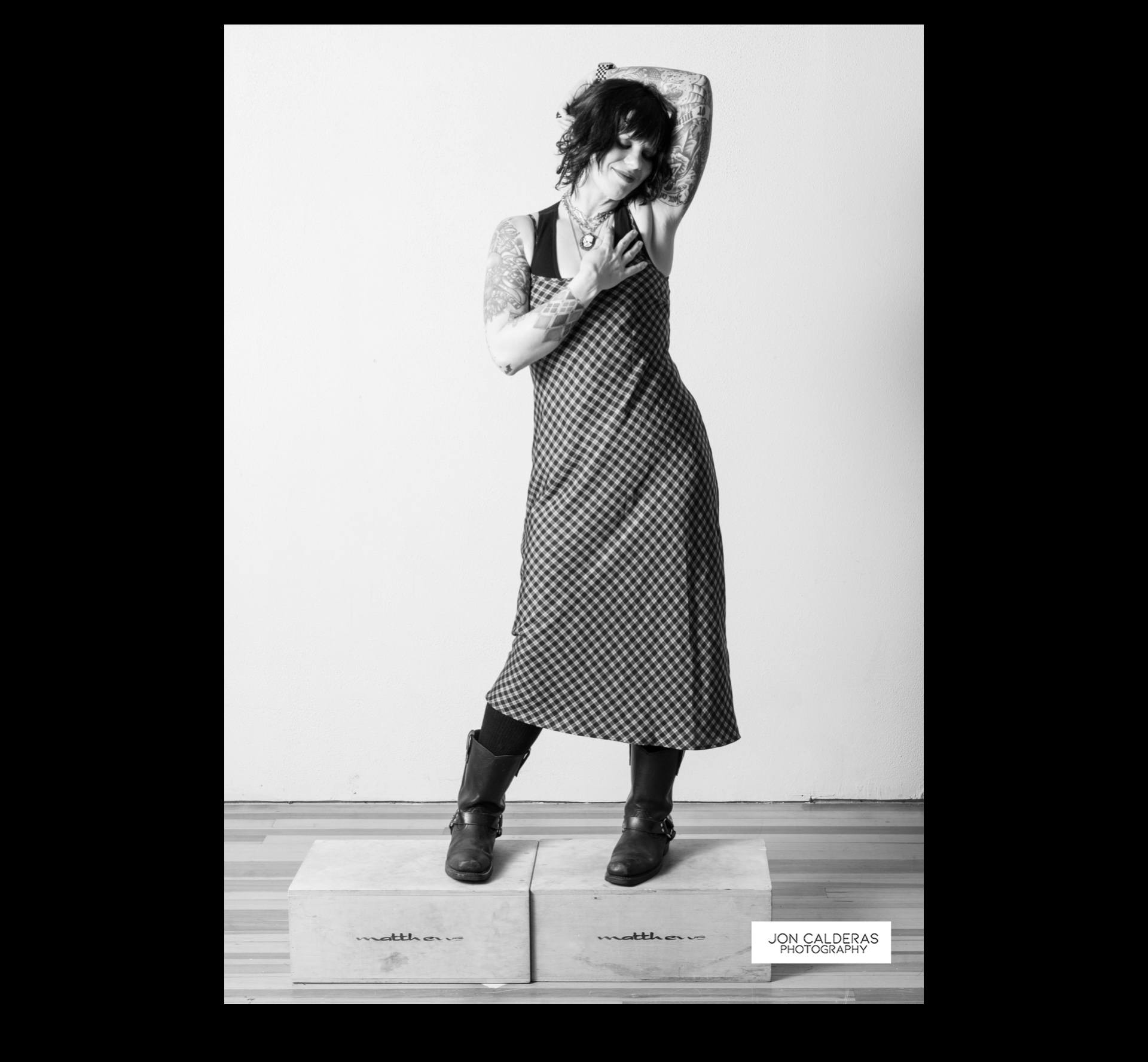 Person with curly hair in a patterned dress and boots, leaning against a wall, standing on a platform, in black and white.