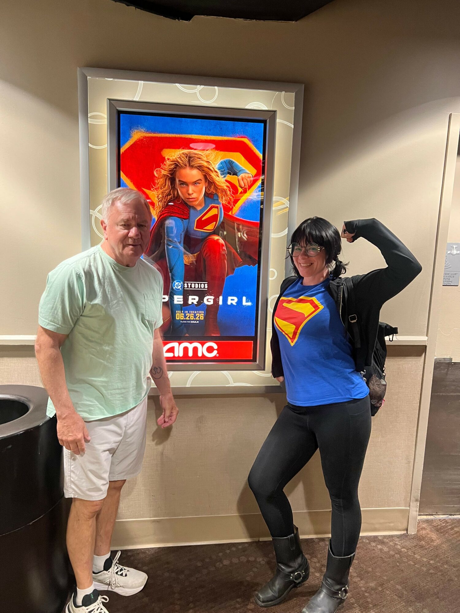 Two people standing in front of a movie poster, one wearing a Superman shirt, the other in a light green shirt and shorts.