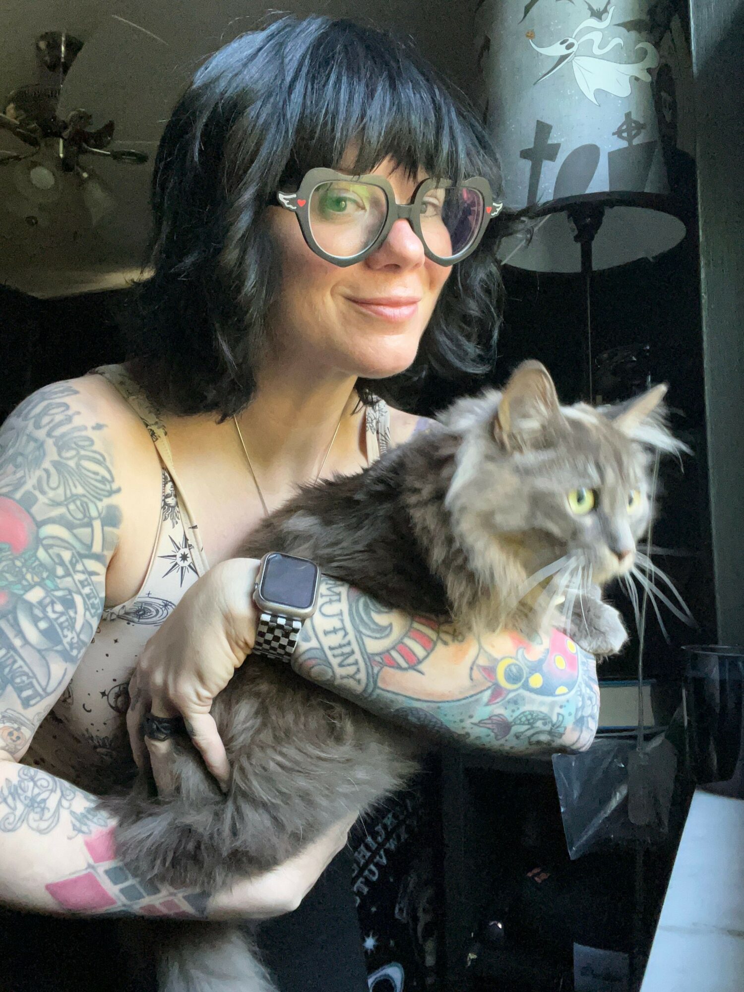 Person with glasses holding a cat, both looking upward, tattoos visible on arms, in an indoor setting.
