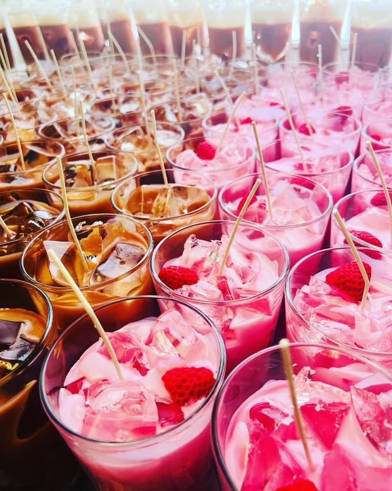 Rows of pink and brown desserts in glasses with skewers, garnished with raspberries, arranged on a table.