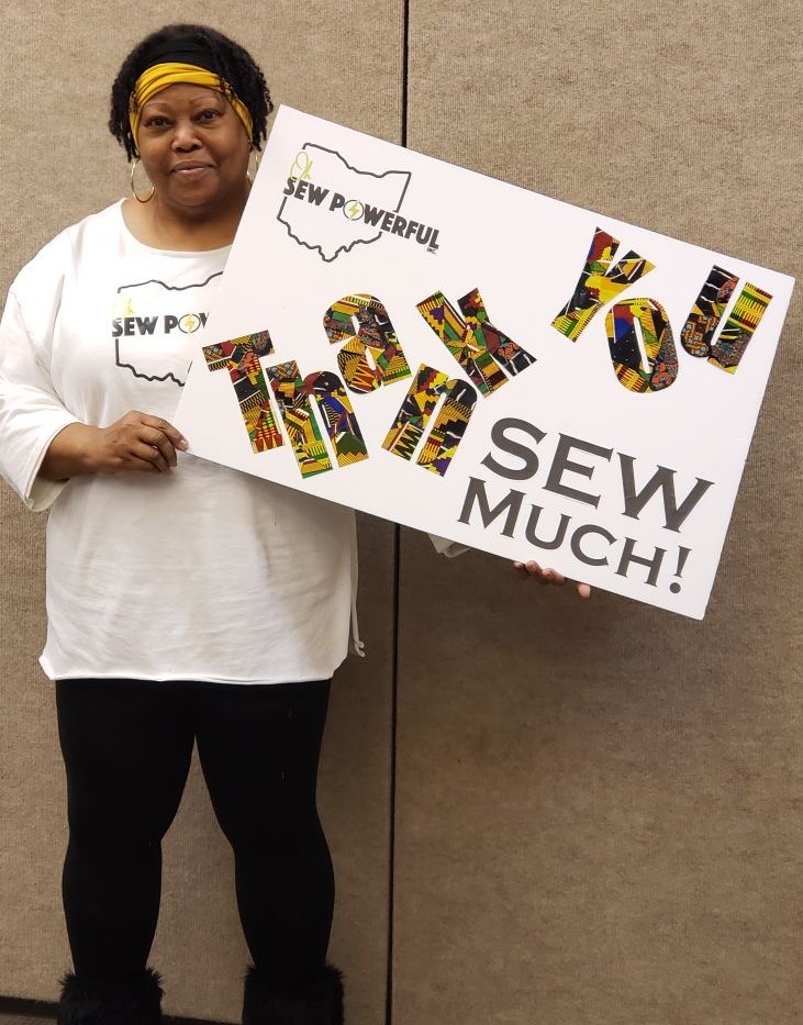 Woman holding a sign with colorful text saying 'Thank You' and 'Sew Much!'.