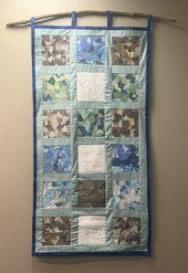 Vertical quilt with patchwork squares in blue, green, brown, and white, hanging from a wooden rod on a wall.