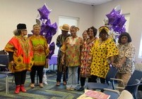 Group of people standing indoors with purple balloons, wearing colorful clothing, some with hats, in a room with chairs and a window.