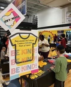 People gather around tables with yellow T-shirts at an indoor event, with a sign promoting T-shirt reuse and kindness.